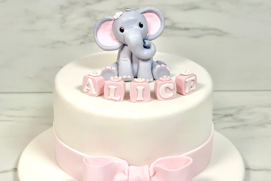 White cake with a pink bow and an elephant decoration on top, and letter blocks for name
